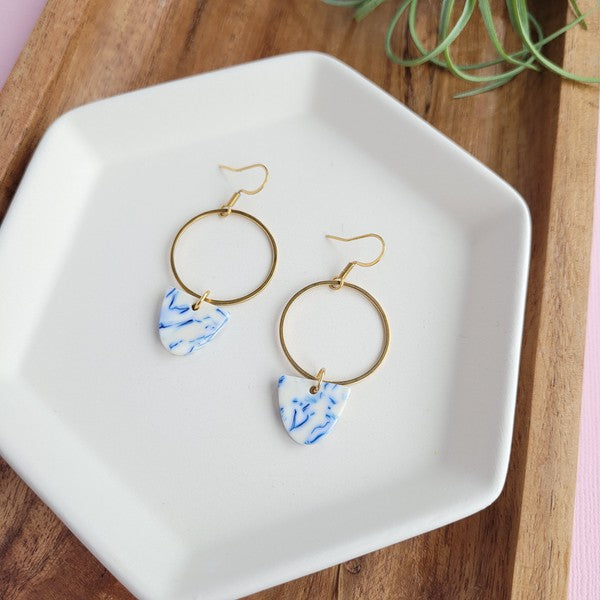 From the Greek Goddess collection, the Iris Earrings - Greek Goddess Blue feature gold hoops and heart-shaped blue and white pendants, elegantly displayed on a hexagonal white dish atop a wooden surface. Made from hypoallergenic stainless steel, they combine both style and comfort seamlessly.