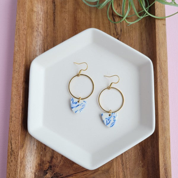 From the Greek Goddess collection, the Iris Earrings - Greek Goddess Blue feature gold hoops and heart-shaped blue and white pendants, elegantly displayed on a hexagonal white dish atop a wooden surface. Made from hypoallergenic stainless steel, they combine both style and comfort seamlessly.