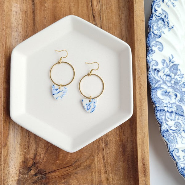 From the Greek Goddess collection, the Iris Earrings - Greek Goddess Blue feature gold hoops and heart-shaped blue and white pendants, elegantly displayed on a hexagonal white dish atop a wooden surface. Made from hypoallergenic stainless steel, they combine both style and comfort seamlessly.