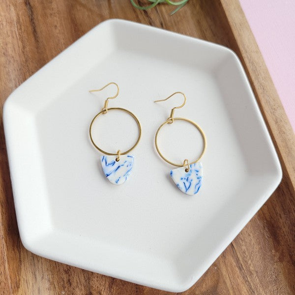 From the Greek Goddess collection, the Iris Earrings - Greek Goddess Blue feature gold hoops and heart-shaped blue and white pendants, elegantly displayed on a hexagonal white dish atop a wooden surface. Made from hypoallergenic stainless steel, they combine both style and comfort seamlessly.