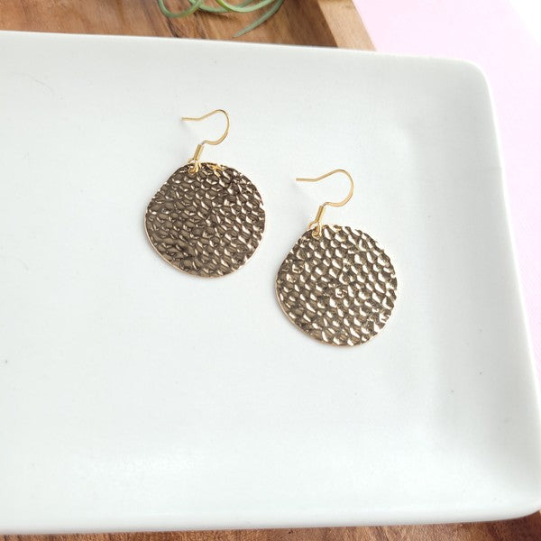 Wearing a black sweater, a person with a cheerful smile adjusts their hair while showcasing the Lucia Earrings - Gold.