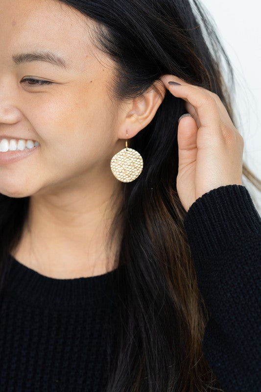 Wearing a black sweater, a person with a cheerful smile adjusts their hair while showcasing the Lucia Earrings - Gold.