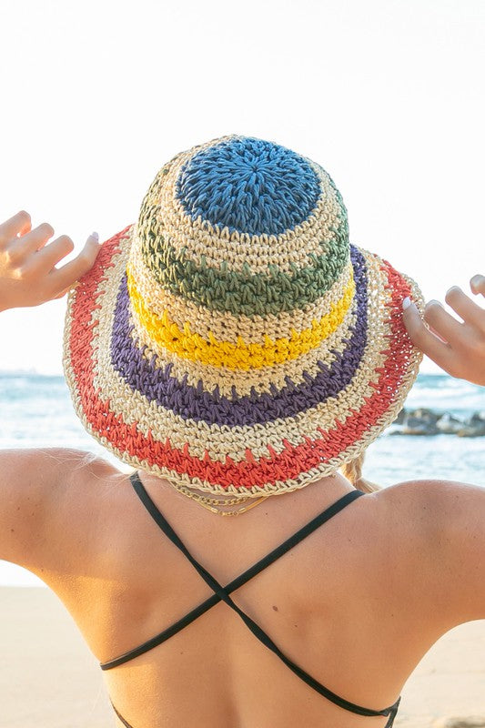 A person from behind wearing a Rainbow Woven Straw Bucket Hat and a black criss-cross strapped swim top at the beach.