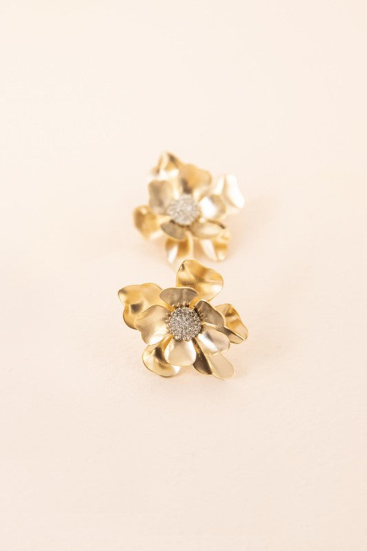 Anemone Post Earrings showcase flower-shaped designs with textured centers and gleaming gold petals, beautifully set against a light beige backdrop.