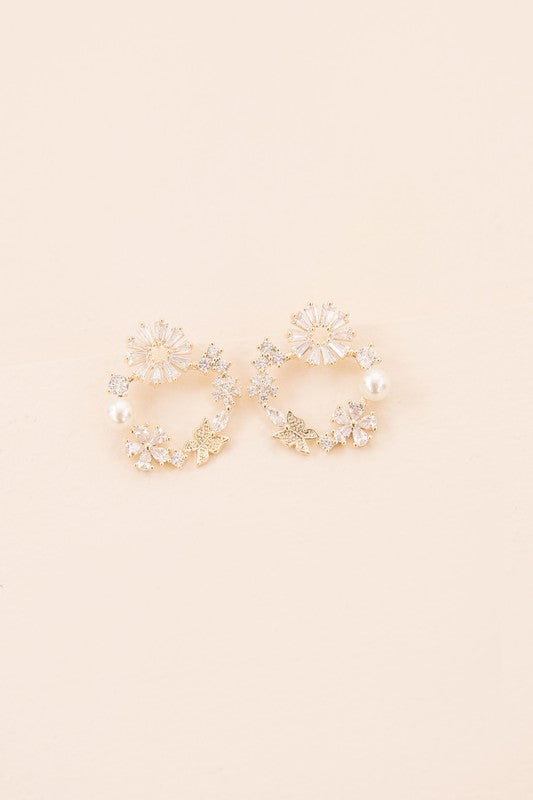 Fable Post Earrings showcase a circular arrangement of gold floral designs and pearl accents, all set against a beige background with a touch of subtle gold plating to enhance their elegance.