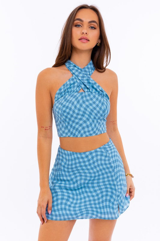 A person stands against a plain background, wearing a chic Halter Neck Crop Top and matching skirt in a checkered pattern.