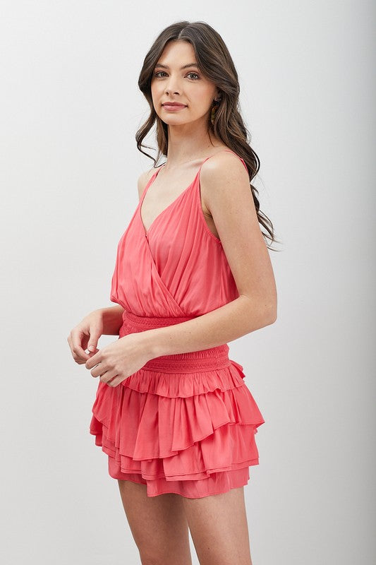 A woman poses against a neutral background wearing the Wrap Smocked Waist Ruffle Dress in coral pink, which features a flattering silhouette with a sleeveless top.