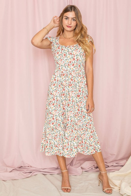 A woman wearing the Flutter Sleeve Ditsy Floral Sundress poses gracefully in front of a pink curtain, her hand gently touching her hair.