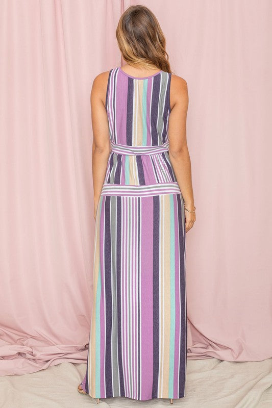 A person wearing the Sleeveless Mix Stripe Maxi Dress, featuring vertical stripes in shades of pink, purple, and blue, stands in front of a beige curtain. This stunning dress is a true testament to elegance and style.