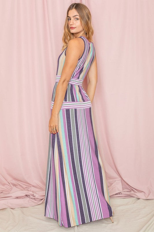 A person wearing the Sleeveless Mix Stripe Maxi Dress, featuring vertical stripes in shades of pink, purple, and blue, stands in front of a beige curtain. This stunning dress is a true testament to elegance and style.