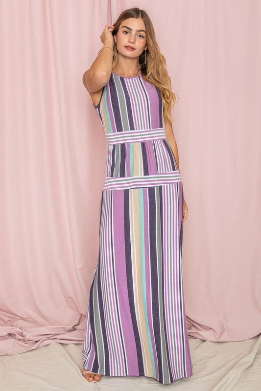 A person wearing the Sleeveless Mix Stripe Maxi Dress, featuring vertical stripes in shades of pink, purple, and blue, stands in front of a beige curtain. This stunning dress is a true testament to elegance and style.