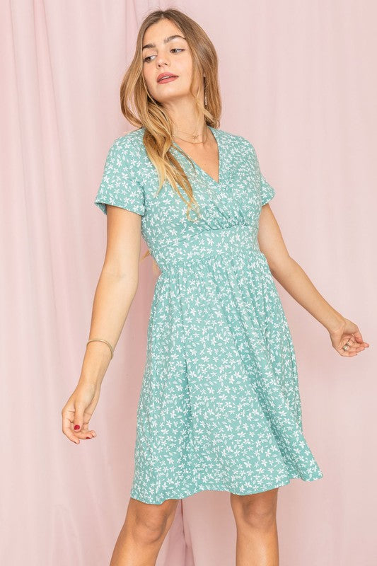 Wearing a Surplice Ditsy Floral Midi Dress, a woman stands against a light pink background, gazing at the camera with a neutral expression. Made from polyester spandex and crafted in the United States, the dress enhances her poised appearance with understated elegance.