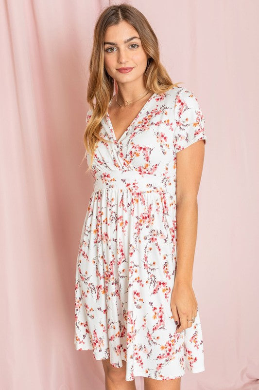 Wearing a Surplice Ditsy Floral Midi Dress, a woman stands against a light pink background, gazing at the camera with a neutral expression. Made from polyester spandex and crafted in the United States, the dress enhances her poised appearance with understated elegance.