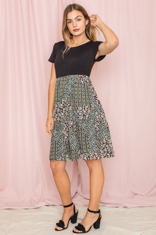 A woman showcases the Floral Mix Print Midi Dress, an ivory midi dress with floral patterns in polyester fabric made in the United States, paired with a short-sleeved black top. She poses against a pink curtain backdrop, with her right hand touching her hair.