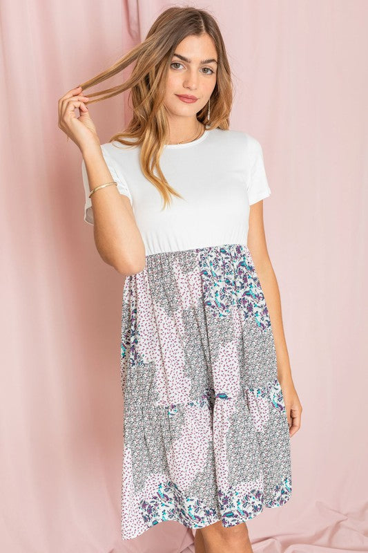 A woman showcases the Floral Mix Print Midi Dress, an ivory midi dress with floral patterns in polyester fabric made in the United States, paired with a short-sleeved black top. She poses against a pink curtain backdrop, with her right hand touching her hair.