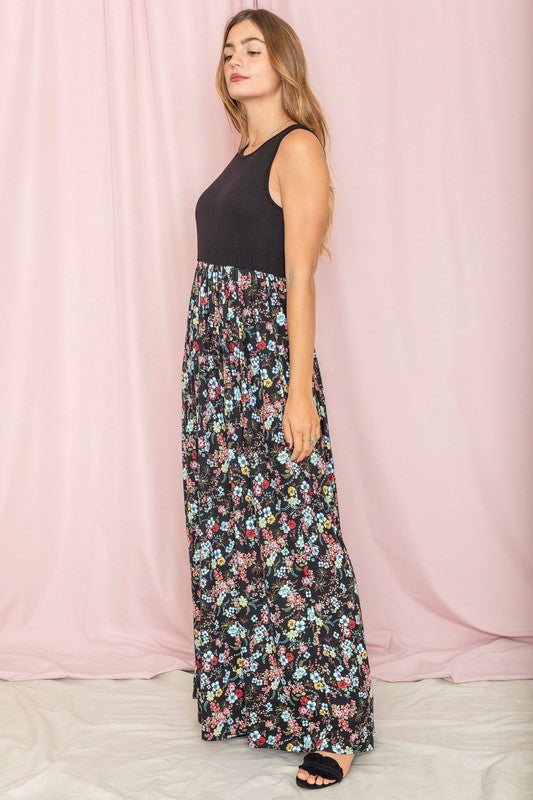 A woman stands against a pink background wearing the Sleeveless Floral Maxi Dress, elegantly paired with a black sleeveless top.