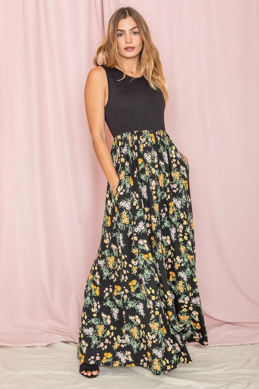 A woman stands against a pink background wearing the Sleeveless Floral Maxi Dress, elegantly paired with a black sleeveless top.