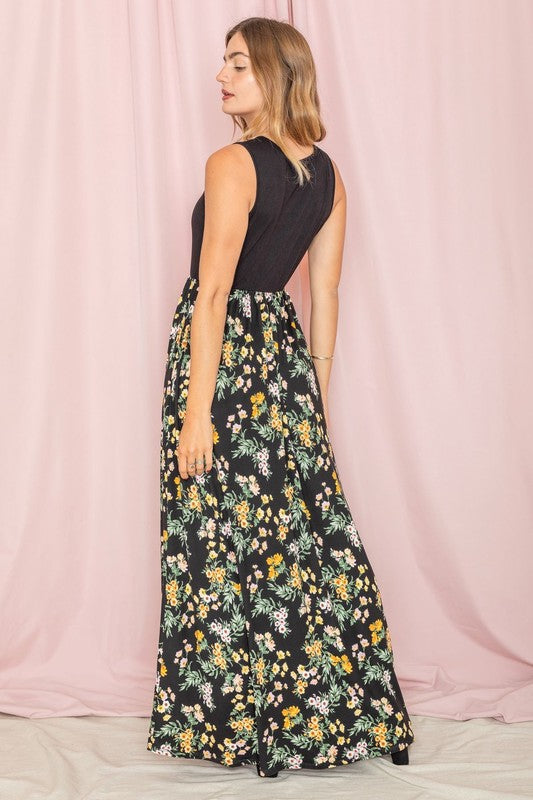 A woman stands against a pink background wearing the Sleeveless Floral Maxi Dress, elegantly paired with a black sleeveless top.