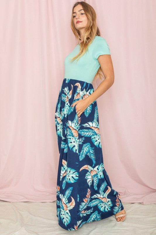 A woman in the Tropical Leaf Short Sleeve Maxi Dress, featuring a blue and green design with a tropical leaf pattern, stands gracefully in front of a pink curtain.