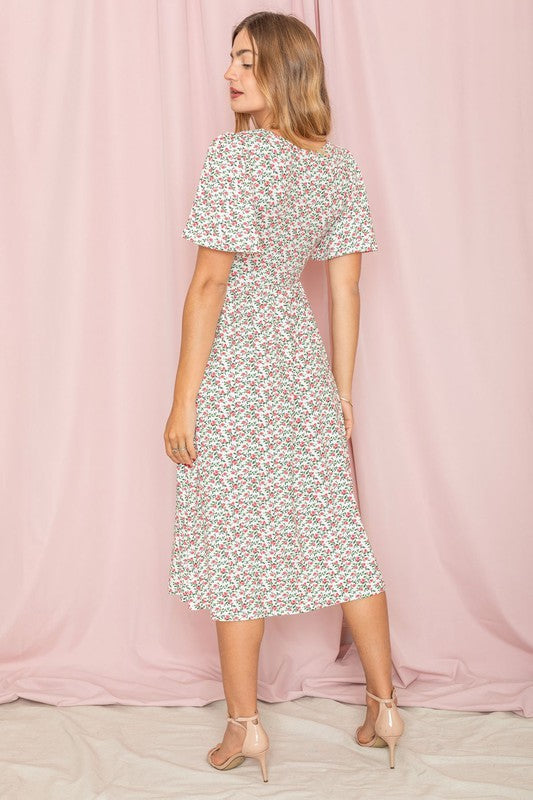 A woman elegantly wearing the Kimono Sleeve Floral Tea Length Dress and black heels stands gracefully in front of a pink curtain.