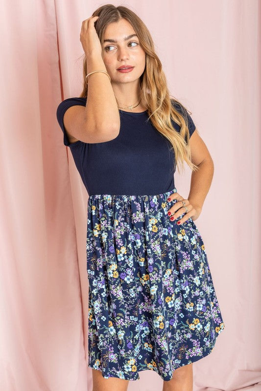 A woman elegantly poses against a pink draped backdrop, dressed in the navy Short Sleeve Ditsy Wildflowers Dress, crafted with pride in the United States.