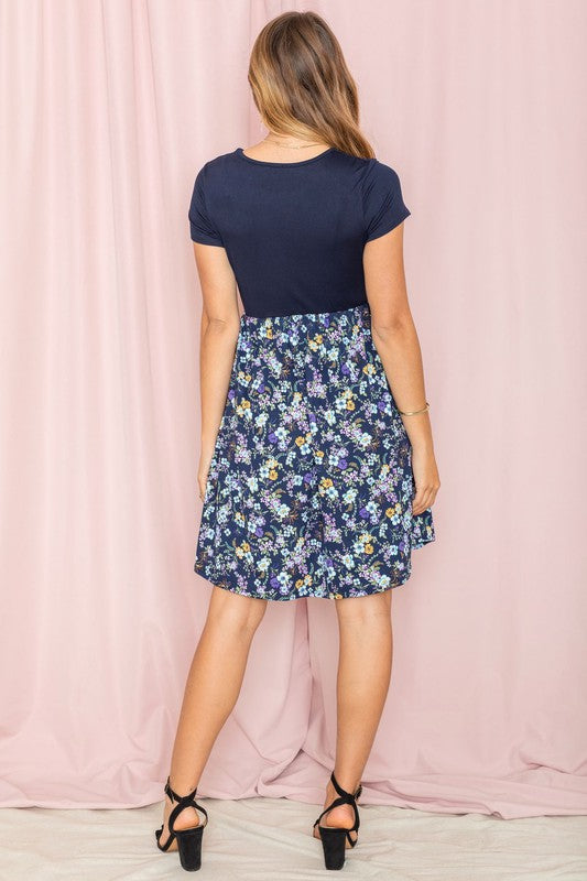 A woman elegantly poses against a pink draped backdrop, dressed in the navy Short Sleeve Ditsy Wildflowers Dress, crafted with pride in the United States.