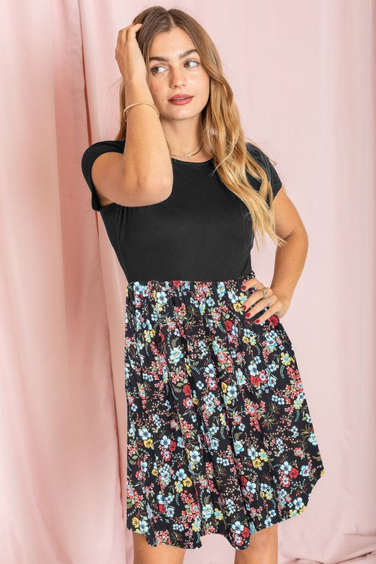 A woman stands gracefully against a pink curtain backdrop, her hand gently touching her hair. She is elegantly showcasing the Short Sleeve Ditsy Wildflowers Dress, made in the United States.