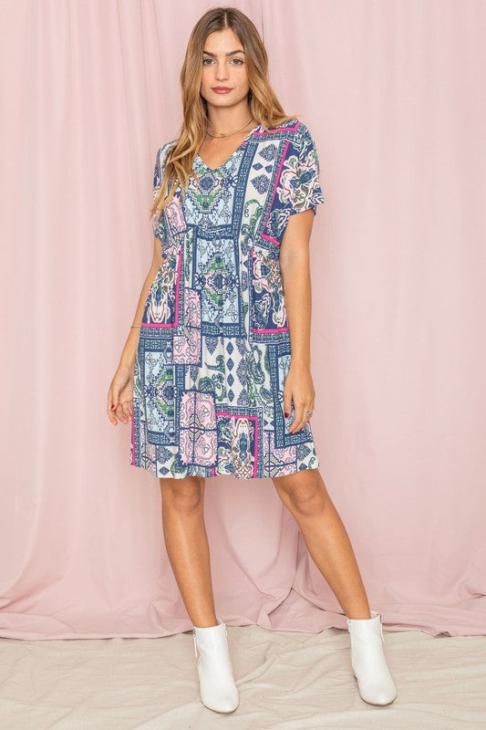 A woman wearing the Patchwork V-Neck Dolman Sleeve Dress and white ankle boots stands gracefully in front of a pink backdrop.