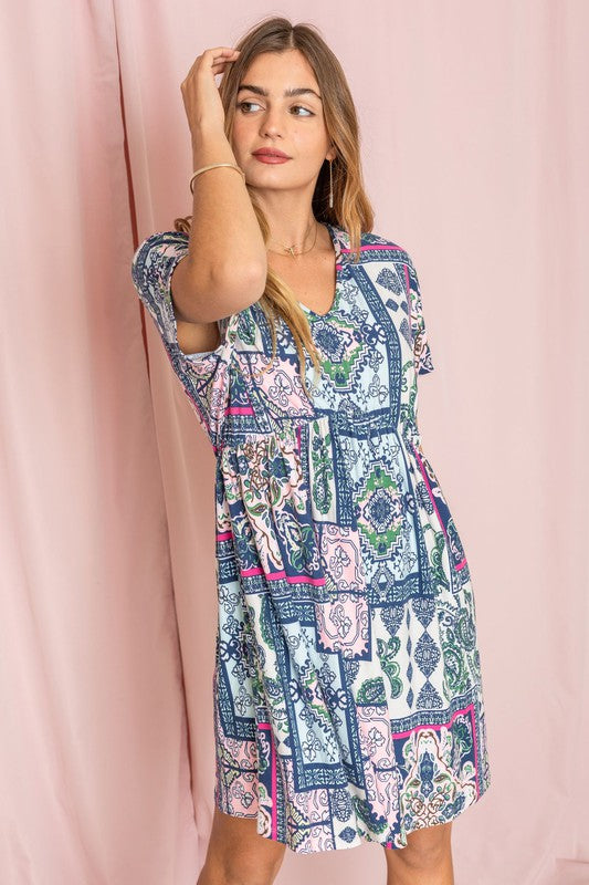 A person wearing a vibrant Patchwork V-Neck Dolman Sleeve Dress featuring blue, pink, and green designs stands in front of a pink curtain.