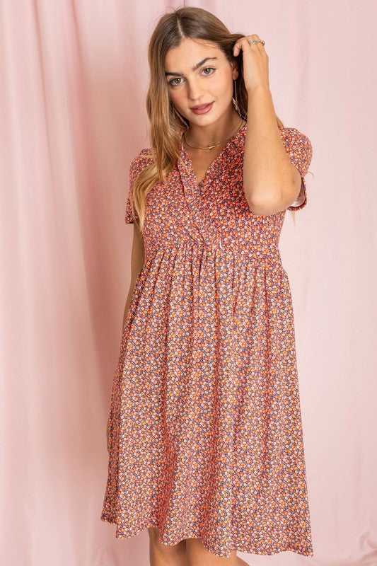 A woman wearing the Floral Surplice Ruffle Hem Dress, crafted in the United States, stands against a pink background, gently touching her hair.