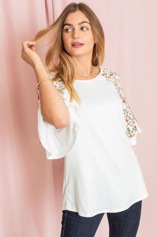 Against a pink backdrop, a woman gracefully poses in the Plus Double Ruffle Sleeve Floral Tunic, featuring a white floral pattern and elegant double ruffle sleeves, crafted with style and elegance in the United States.