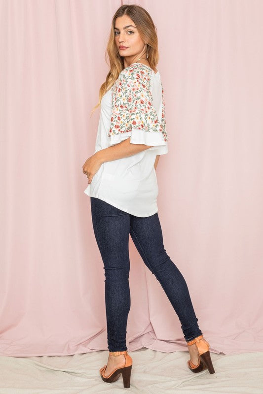 Against a pink backdrop, a woman gracefully poses in the Plus Double Ruffle Sleeve Floral Tunic, featuring a white floral pattern and elegant double ruffle sleeves, crafted with style and elegance in the United States.