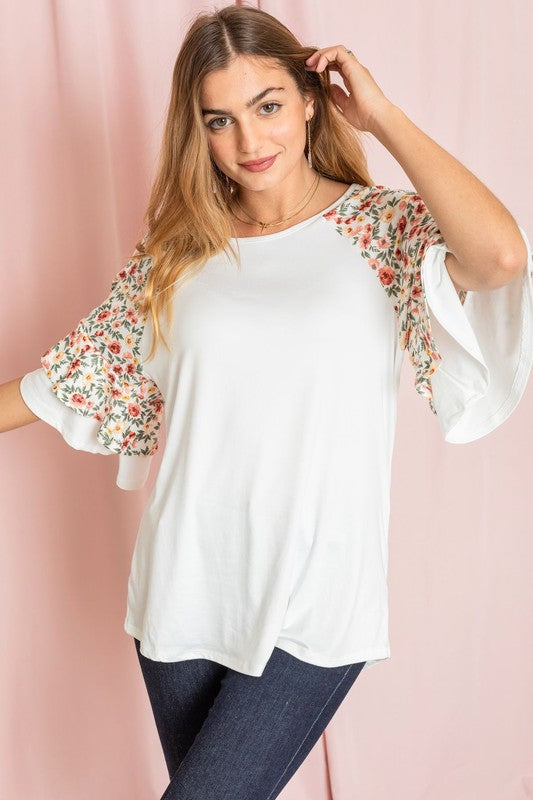 Against a pink backdrop, a woman gracefully poses in the Plus Double Ruffle Sleeve Floral Tunic, featuring a white floral pattern and elegant double ruffle sleeves, crafted with style and elegance in the United States.