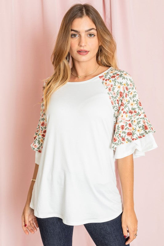 Against a pink backdrop, a woman gracefully poses in the Plus Double Ruffle Sleeve Floral Tunic, featuring a white floral pattern and elegant double ruffle sleeves, crafted with style and elegance in the United States.