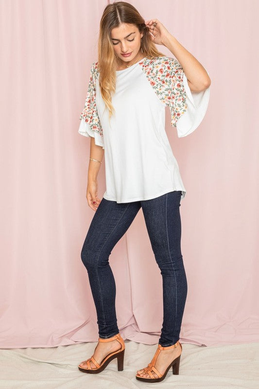 Against a pink backdrop, a woman gracefully poses in the Plus Double Ruffle Sleeve Floral Tunic, featuring a white floral pattern and elegant double ruffle sleeves, crafted with style and elegance in the United States.