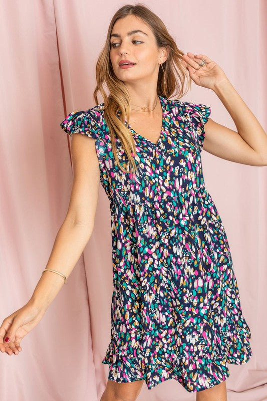 A person stands against a pink background wearing the Colorful Abstract Flutter Sleeve Mini Dress paired with white ankle boots.