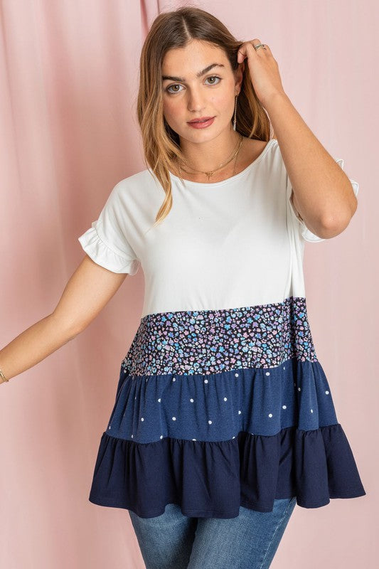 Against a pink backdrop, a woman showcases the Frill Sleeve Tiered Color Block Tunic, characterized by its floral and polka dot ruffles. She pairs it elegantly with blue jeans, creating an ensemble that exudes charm and captures the essence of playful sophistication.