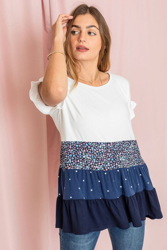 Against a pink backdrop, a woman showcases the Frill Sleeve Tiered Color Block Tunic, characterized by its floral and polka dot ruffles. She pairs it elegantly with blue jeans, creating an ensemble that exudes charm and captures the essence of playful sophistication.