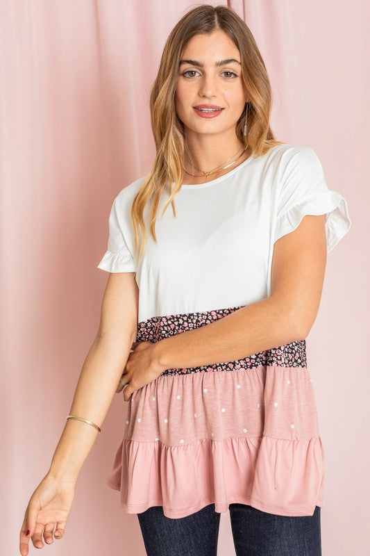 Against a pink backdrop, a woman showcases the Frill Sleeve Tiered Color Block Tunic, characterized by its floral and polka dot ruffles. She pairs it elegantly with blue jeans, creating an ensemble that exudes charm and captures the essence of playful sophistication.