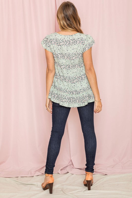 A woman in a Plus Ditsy Floral Ruffle Hem Blouse and jeans stands gracefully in front of a beige curtain, her outfit exuding charm and casual elegance.