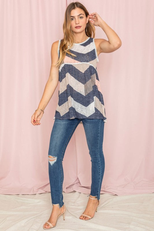 A woman with long hair is wearing the Pom Pom Chevron Top, crafted from rayon and polyester, paired with jeans and standing against a pink background.