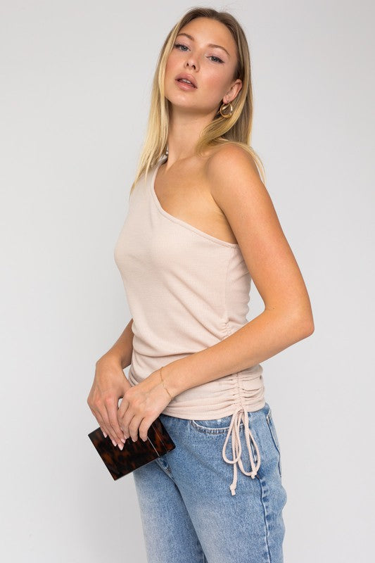 A person with long hair is elegantly styled in a fashion ensemble featuring a One Shoulder Side Rushing Top and jeans, holding a small rectangular object.