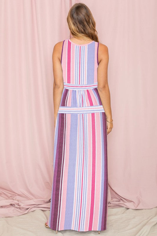 A person wearing the Sleeveless Mix Stripe Maxi Dress, featuring vertical stripes in shades of pink, purple, and blue, stands in front of a beige curtain. This stunning dress is a true testament to elegance and style.