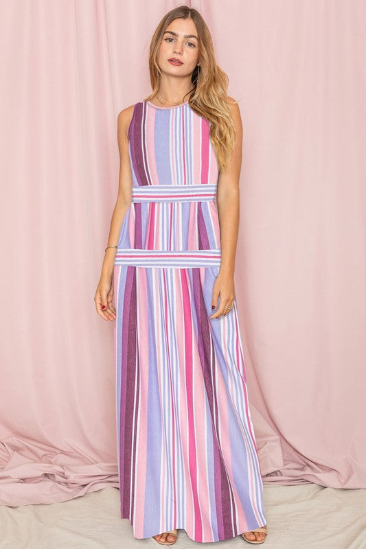 A person wearing the Sleeveless Mix Stripe Maxi Dress, featuring vertical stripes in shades of pink, purple, and blue, stands in front of a beige curtain. This stunning dress is a true testament to elegance and style.