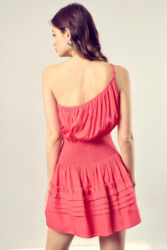 A woman in a Pleated Detail One Shoulder Cami Dress featuring a sleeveless, coral design with a cinched waist and tiered, pleated skirt.