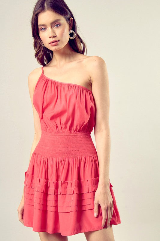 A woman in a Pleated Detail One Shoulder Cami Dress featuring a sleeveless, coral design with a cinched waist and tiered, pleated skirt.