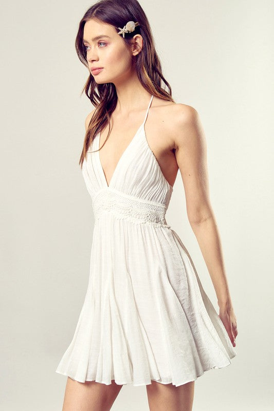 A person dressed in the Lace Trim with Back Drawstring Dress, featuring a sleeveless design, a white pleated skirt, and intricate lace trim, gazes to the side against a neutral backdrop.