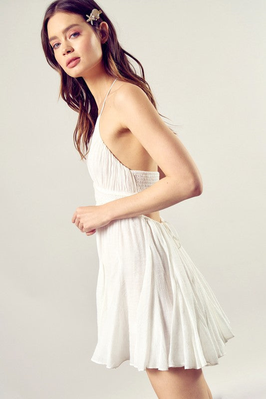 A person dressed in the Lace Trim with Back Drawstring Dress, featuring a sleeveless design, a white pleated skirt, and intricate lace trim, gazes to the side against a neutral backdrop.