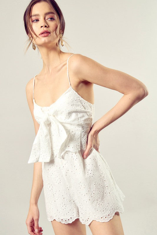 A woman wearing the Scallop Edge Front Tie-Up Romper in white, featuring a sleeveless design and eyelet texture, stands against a plain background, radiating boho style charm.