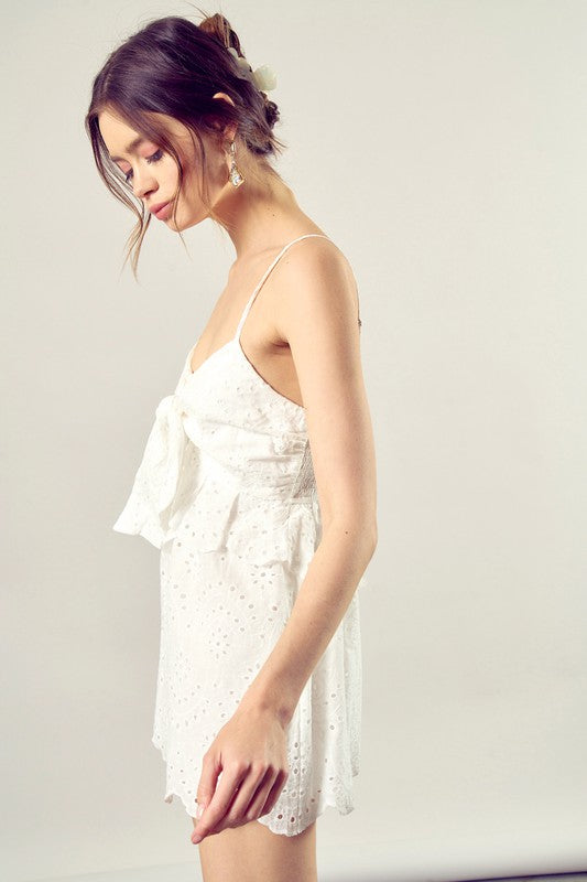 A woman wearing the Scallop Edge Front Tie-Up Romper in white, featuring a sleeveless design and eyelet texture, stands against a plain background, radiating boho style charm.